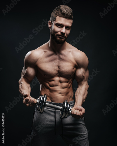 Muscular hunk with dumbbell posing in studio. Fitness male model holding gym equipment. Shirtless man at black background.