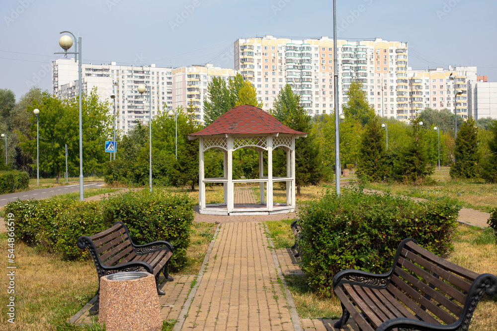 Summer city park, paved path and wooden gazebo against the backdrop of residential buildings Summer city park, paved path and wooden gazebo against the backdrop of residential buildings