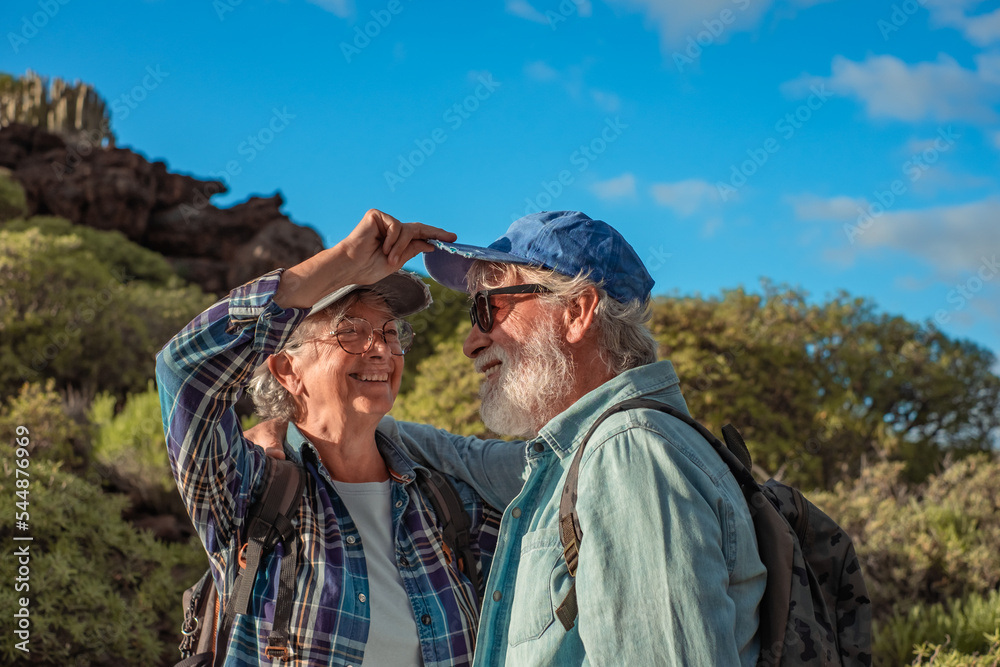 Lovely happy senior couple hugging in mountain trekking enjoying nature, freedom and healthy lifestyle. Smiling old retirees in hat and casual clothes among green bushes and blue sky