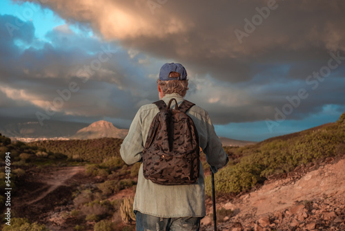 Back view of active senior man enjoying trekking at sunset light. Elderly caucasian male with hat and backpack walking in mountain with a stick. Dramatic sky