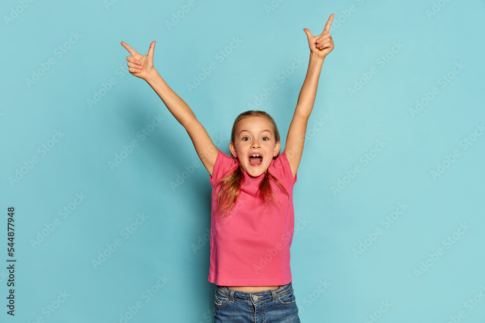 Portrait of little beautiful girl, child in pink T-shirt posing isolated over blue studio background. Winning look. Happy - obrazy, fototapety, plakaty 