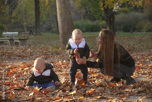 A mother walks her twin children with the help of her daughter who walks her other brother. They hold the children's hands and make sure they don't fall. In the park in late fall, the park is littered