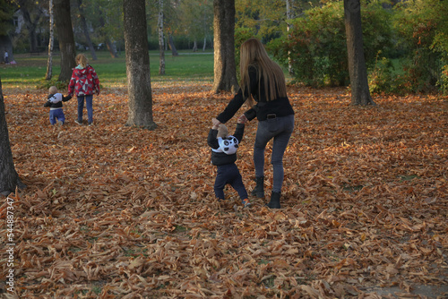 A mother walks her twin children with the help of her daughter who walks her other brother. They hold the children's hands and make sure they don't fall. In the park in late fall, the park is littered