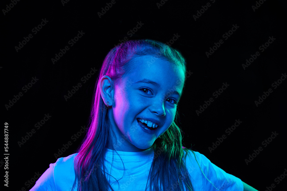 Portrait of little beautiful girl, child in white T-shirt posing, smiling isolated over black studio background in neon light