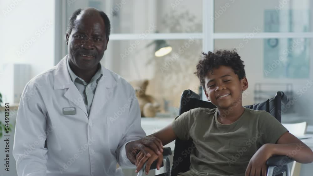 Portrait of African American doctor and boy in wheelchair holding hands and posing for camera in pediatric clinic