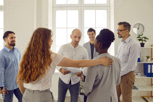 Corporative business training in office with people standing in round and listening man colleague. Man talking to an african american woman and the woman pats her on shoulder. Team building seminar.