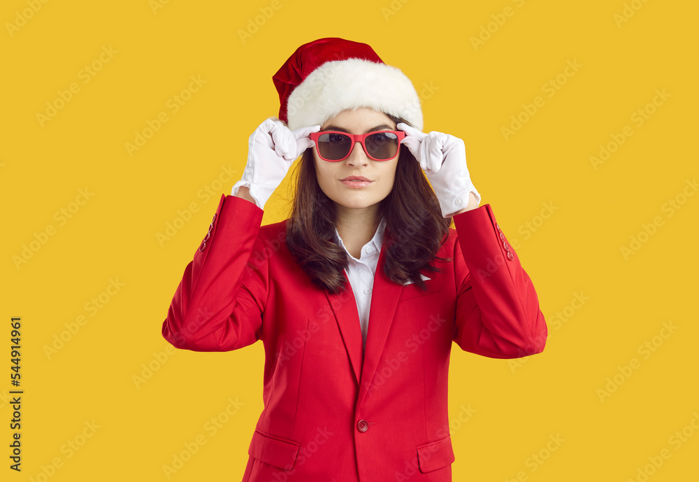 Studio shot of young woman in stylish Santa Christmas costume. Confident unsmiling model wearing trendy red suit, Santa hat, white gloves and sunglasses standing isolated on yellow background