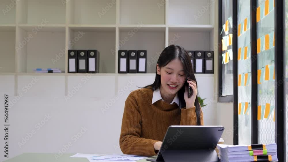 Young busy asian business woman working on laptop talking on the phone. Ethnic female manager making business call on mobile in office. Young busy asian business woman working on laptop talking on the phone. Ethnic female manager making business call on mobile in office.