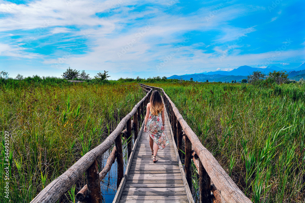 A girl walking in a wooden walkways in the middle of the Massaciuccoli lake, Tuscany, Italy on a sunny day. With nature and fauna all around.