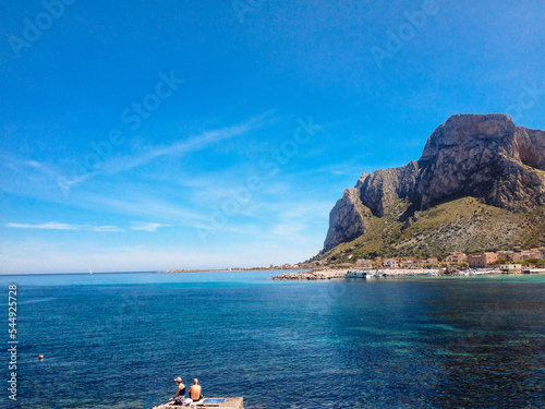 Sferracavallo, Province of Palermo, Sicily, Italy. With rocky coast and blue sea, coastal stones and people. Here on a sunny day with mountain in the background.