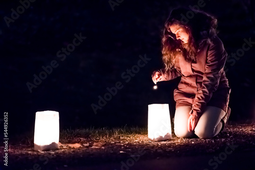 One Young Woman lights up with match, stick Christmas Eve candle light, lantern in paper bags at dark night, evening outside, outdoors on street, road