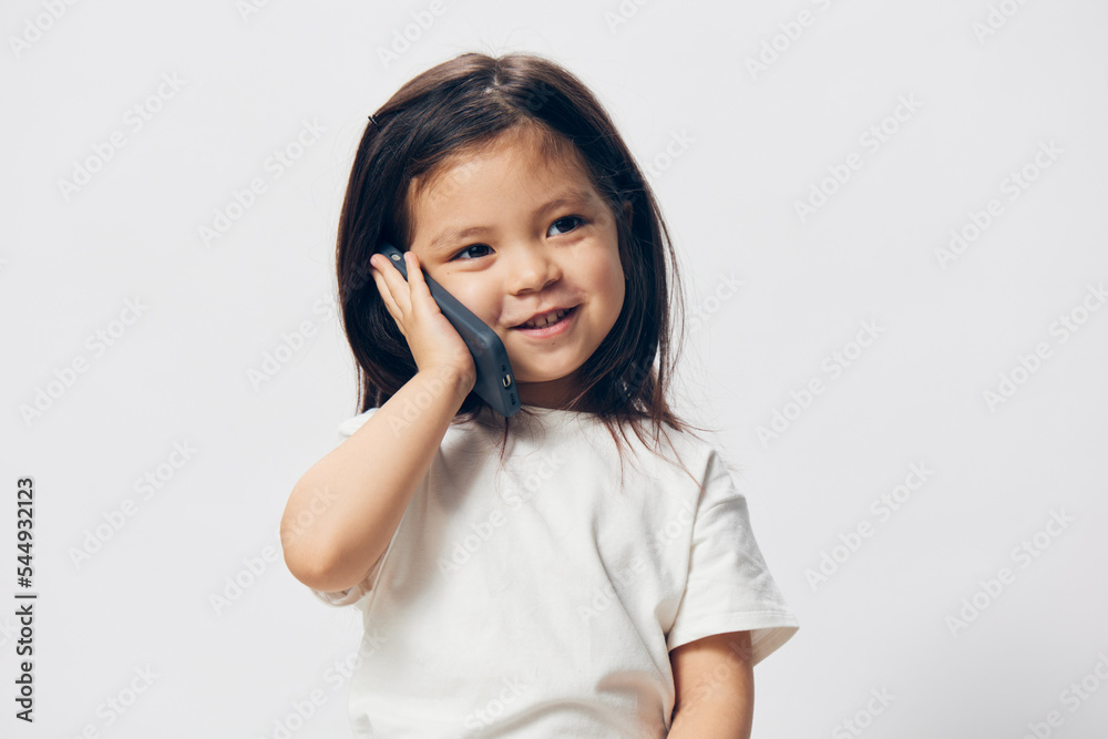 a cute little preschool girl is standing on a white background in a white T - shirt , sitting on a chair and talking on the phone looking away . The topic of children's conversation a cute little preschool girl is standing on a white background in a white T - shirt , sitting on a chair and talking on the phone looking away . The topic of children's conversation