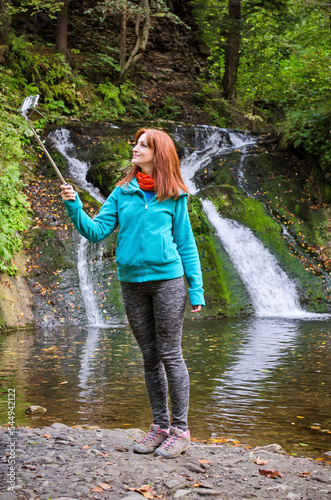 Young red haired woman blogger in autumn mountains near lake and waterfall is recording video, taking photo of herself with selfie stick. She is dressed in blue jacket, orange scarf and gray pants