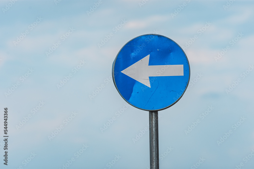 Regulatory road sign to turn left in the first intersection with a white arrow on a blue circle against clear sky