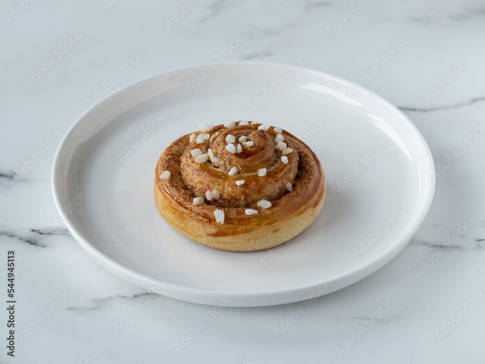Traditional sweden cinnamon bun kanelbullar with cardamom sprinkled with pearl sugar. White plate on a marble table. Homemade pastry. Part of fika Traditional sweden cinnamon bun kanelbullar with cardamom sprinkled with pearl sugar. White plate on a marble table. Homemade pastry. Part of fika