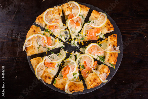 Pizza Gourmet Napoletana con salmone, fette di limone rucola e cipolla croccante tagliata e servita su un piatto nero in una pizzeria napoletana