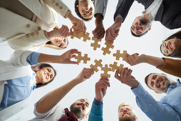 Wooden pieces of puzzles in hands of cheerful and joyful business people who connect them. Smiling people stand in circle with pieces of puzzles that mean their contribution to business development.