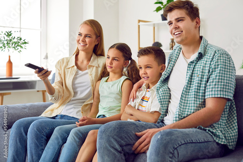 Young cheerful family with children relax watching television programs together. Woman uses remote control to switch channels on TV while sitting on sofa with her husband and small son and daughter.