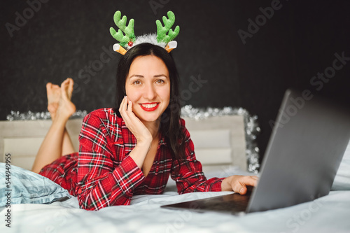 Beautiful excited smiling brunette woman in red pajamas lies in bed in Christmas reindeer horns works with laptop in dark interior bedroom. Young female freelancer at home in happy New Year mood. photo