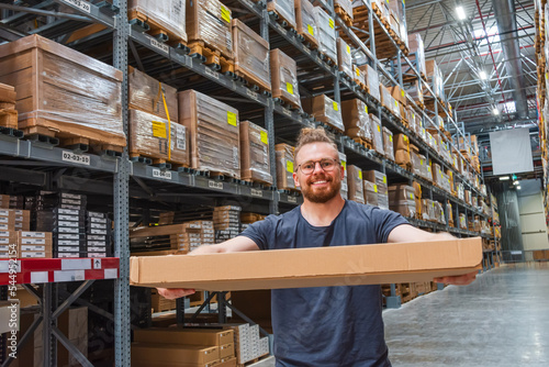 Caucasian young male smiling in glasses is warehouse worker holding and giving box to customer, against the backdrop of an aisle of racks for storage. Assembly preparation for shipment of goods.