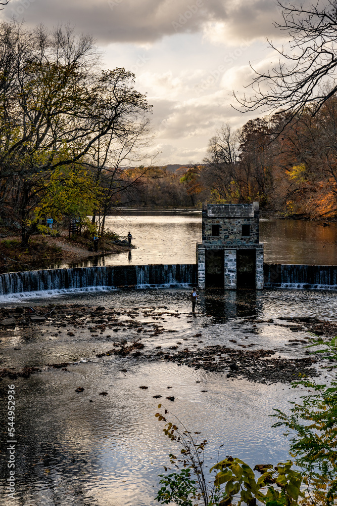 Morristown, NJ - USA - Nov 5, 2022 An autumnal vertical view of New Jersey's historic stone Speedwell Dam during sunset. Fly fisherman in the foreground and people hiking in the distance.