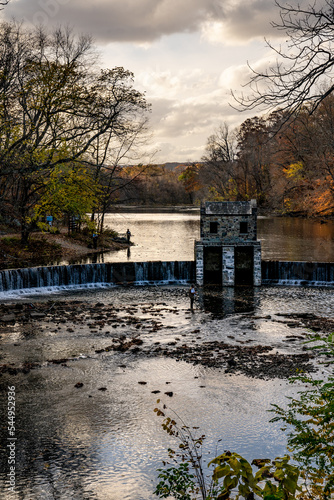 Obraz na płótnie Morristown, NJ - USA - Nov 5, 2022 An autumnal vertical view of New Jersey's historic stone Speedwell Dam during sunset. Fly fisherman in the foreground and people hiking in the distance. w salonie