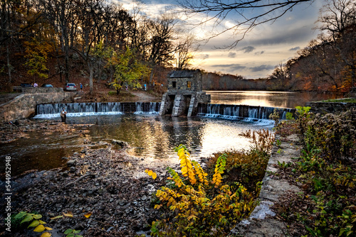 Morristown, NJ - USA - Nov 5, 2022 An autumnal horizontal wide angle view of New Jersey's historic stone Speedwell Dam during sunset. Fly fisherman in the foreground and people hiking in the distance.