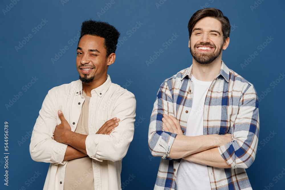 Young two friends cheerful playful happy men 20s wears white casual shirts look camera together hold hands crossed folded wink isolated plain dark royal navy blue background. People lifestyle concept. Young two friends cheerful playful happy men 20s wears white casual shirts look camera together hold hands crossed folded wink isolated plain dark royal navy blue background. People lifestyle concept.