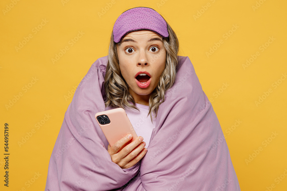 Young shocked woman wear pyjamas jam sleep eye mask wrapped in duvet blanket use mobile cell phone rest relax at home isolated on plain yellow background studio portrait. Good mood night nap concept. Young shocked woman wear pyjamas jam sleep eye mask wrapped in duvet blanket use mobile cell phone rest relax at home isolated on plain yellow background studio portrait. Good mood night nap concept.