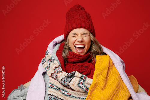 Vászonkép Young sad frozen woman wear scarf hat wrapped in many plaids close eyes scream shout isolated on plain red background studio portrait