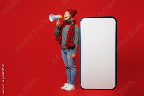 Tablou canvas Full body young woman wear grey sweater scarf hat big area blank screen mobile cell phone scream in megaphone isolated on plain red background
