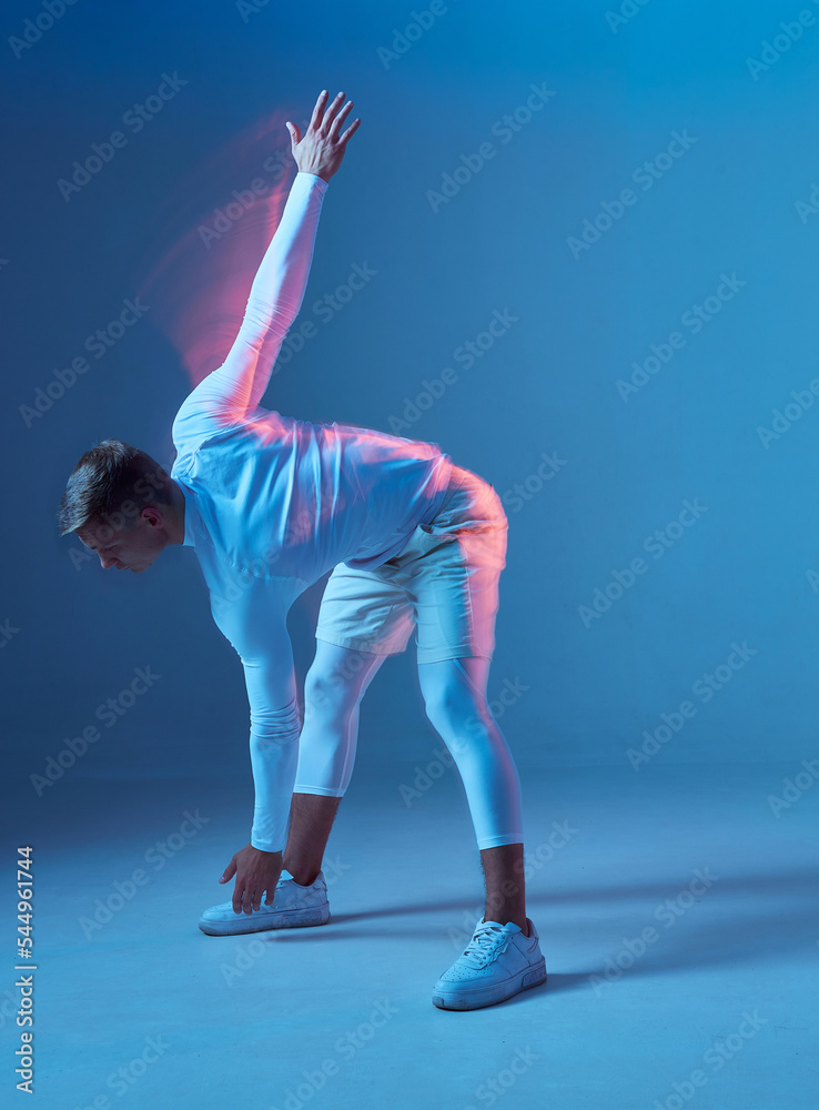 Side view of an athlete doing warm up before workout. Man in white sportswear on blue background does tilt forward.