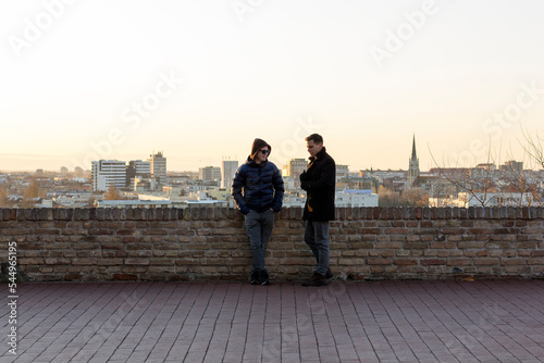Two friends are standing on the terrace of the Petrovaradin fortress talking, having fun, behind them a panoramic view of the city of Novi Sad in the early evening.