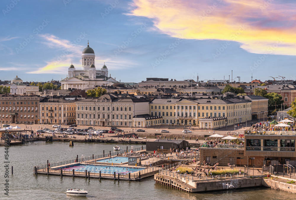 Fototapeta premium Helsinki, Finland - July 20, 2022: The white Cathedral with green domes towers over the port, the public swimming pool, Atlas Restaurant and cityscape under blue sunset cloudscape