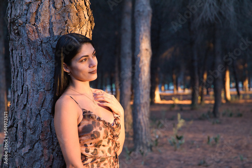 South American woman, young and beautiful, brunette, in leopard dress, leaning on the trunk of a tree, in the golden hour. Concept beauty, fashion, autumn, golden hour, melancholy.