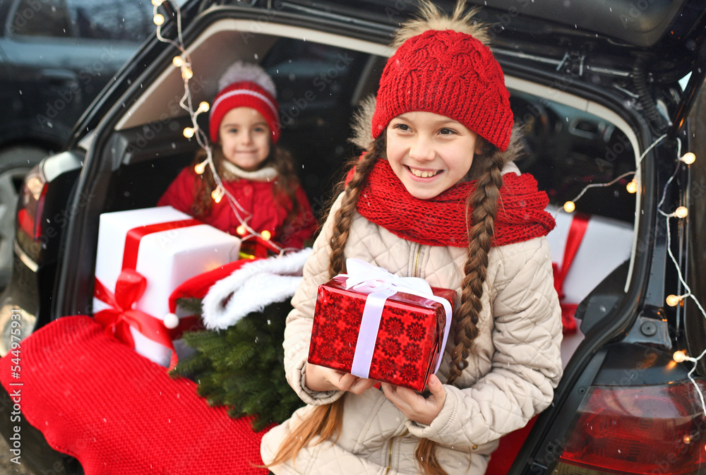 two little girls are smiling while sitting in the trunk of a car and holding Christmas gifts in their hands against the background of Christmas tree branches, a red plaid and Christmas lights.