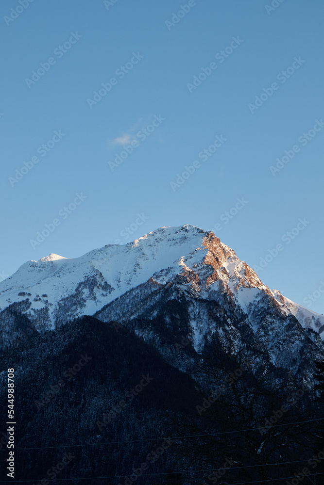 mountain peak at sunset, part of mountain is illuminated by rays of sun, clear sky and coniferous forest below, evening time