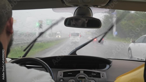 A caucasian bearded man drives a car along the highway when it rains. A man rides in a stream of cars on a city road.