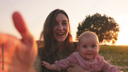Good-looking young Caucasian woman holding lovely toddler in hands, talking selfie on smartphone, looking at camera in nature. Girl and baby smiling. Mother and child concept. Outdoor. Field. Sunset