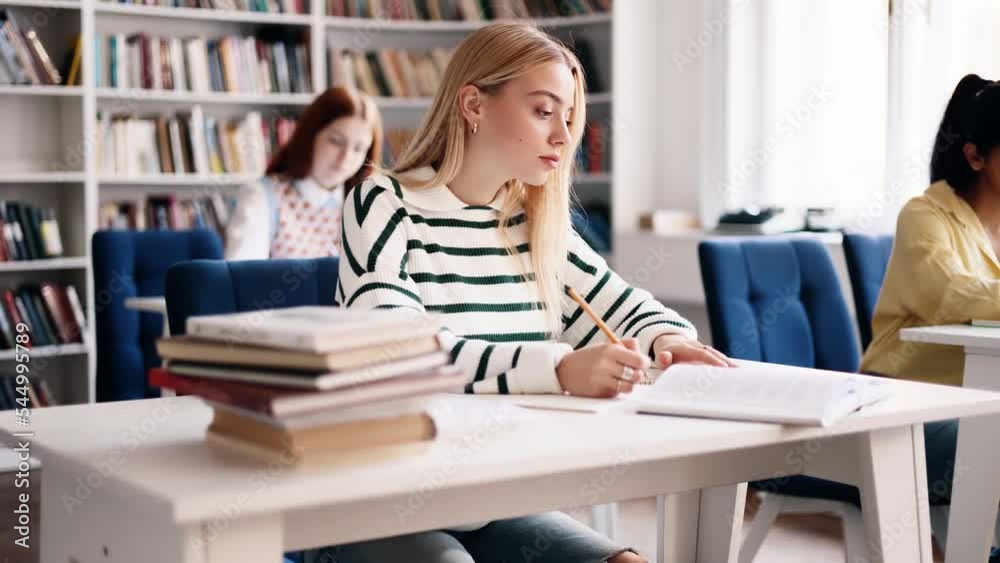 Attractive young blond girl preparing for exam and studying at university library. Charming young student focused on learning and make notes in her notebook in multiethnic group. Attractive young blond girl preparing for exam and studying at university library. Charming young student focused on learning and make notes in her notebook in multiethnic group.
