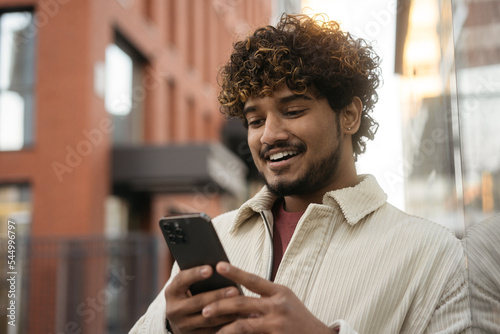 Smiling curly haired Indian man using mobile app shopping online, choosing something on website. Happy asian hipster guy playing mobile game standing on city street. Modern technology concept