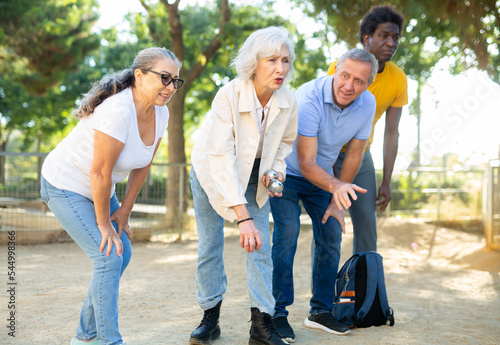 Senior mature white woman preparing to throw a boule ball in petanque game in the park with other people standing beside  © JackF