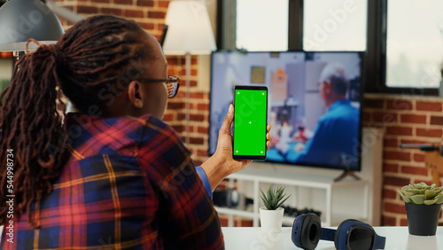 Young manager vertically holding mobile phone with greenscreen display, sitting at home office. Working with isolated chroma key template and blank copyspace, mock up background. Tripod shot.