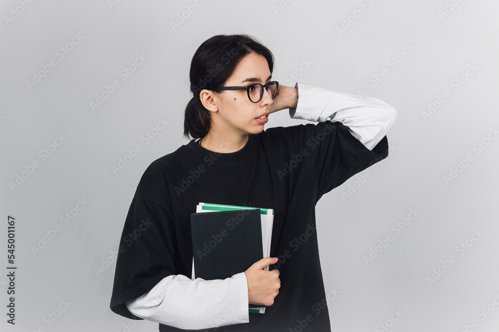 Young student girl with glasses looks away. She holds a stack of books for reading, studying or education