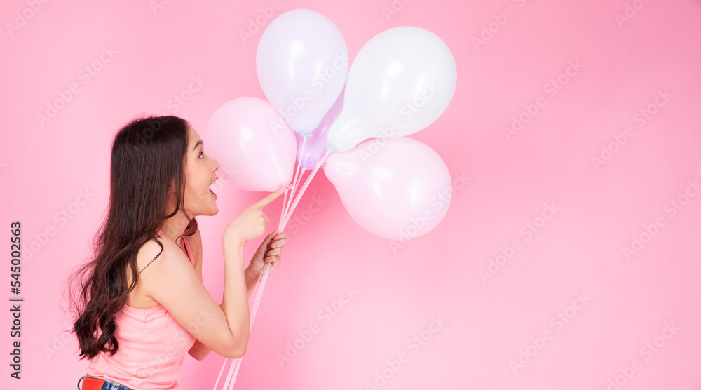 Excited cheerful asian woman holding balloons in hands using finger pointing to copy space standing over isolated pink background. Joyful teenager girl with pastel balloons shocked  amazed expression.