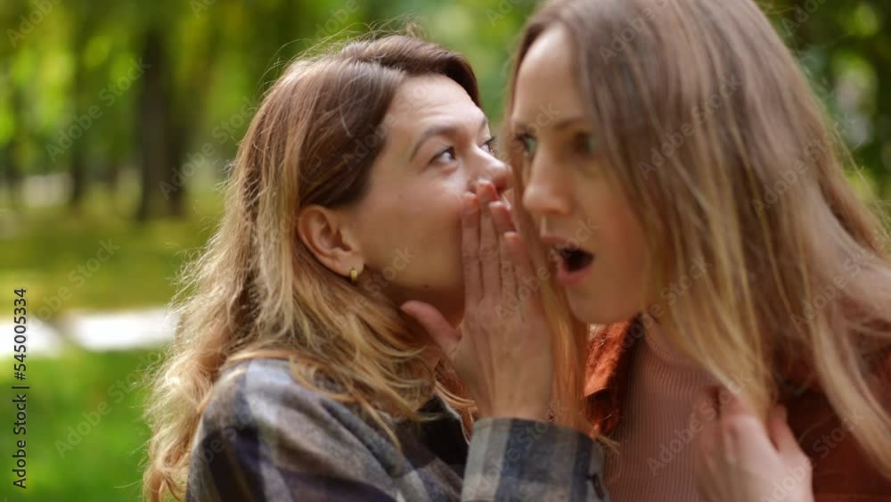 Rack focus from woman whispering on ear to surprised curios friend listening talking. Two young Caucasian beautiful ladies sharing rumors gossiping in sunny park outdoors. Femininity concept Rack focus from woman whispering on ear to surprised curios friend listening talking. Two young Caucasian beautiful ladies sharing rumors gossiping in sunny park outdoors. Femininity concept