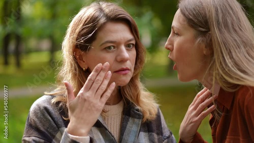 Close-up portrait of shocked surprised Caucasian woman in sunny autumn park with friend appearing whispering rumors on ear. Astonished lady sharing gossip with friend outdoors