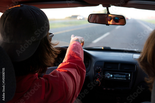young adult man with long hair driving a car on the road and pointing at something.