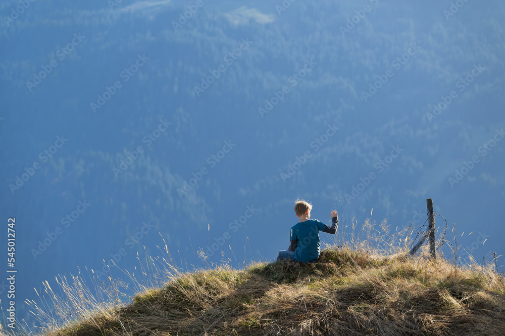 Obraz premium Eleven year old boy sits on a mountains ridge and lets grass seeds fly
