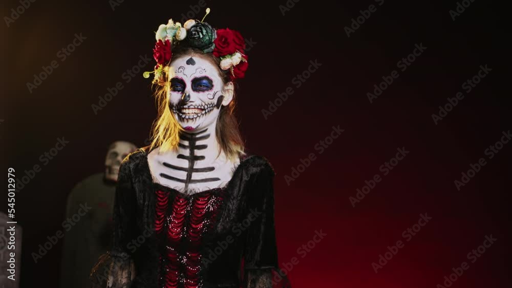 Holy mexican entity laughing and acting scary in studio, looking like la cavalera catrina on day of the dead traditional holiday. Smiling and wearing halloween costume with flowers. Handheld shot.